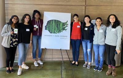 Seven students stand smiling around a poster with the Women in Data Science Berkeley logo. 